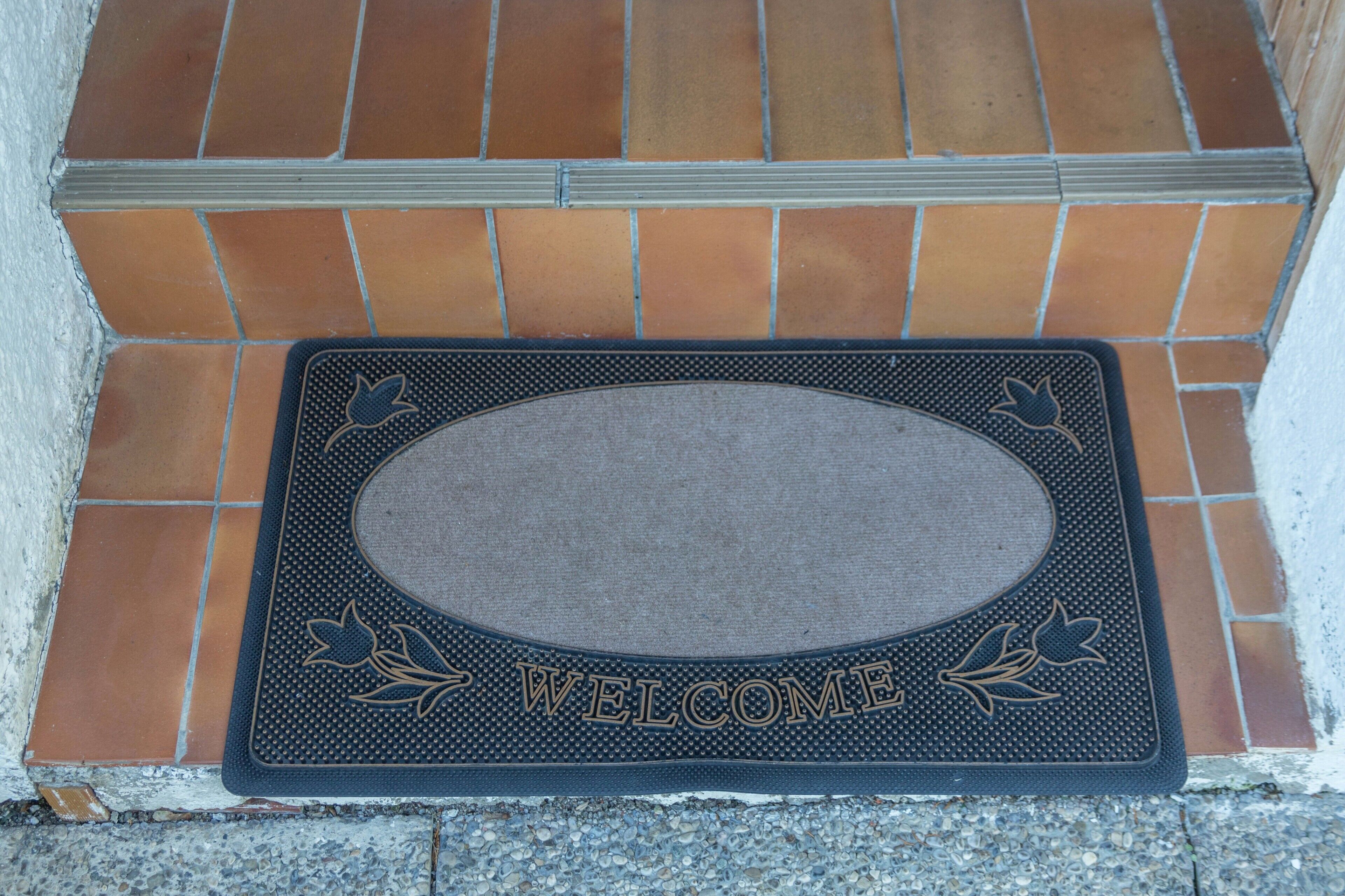 Un paillasson avec l’inscription « BIENVENUE » et des motifs de feuilles, posé sur un sol carrelé.