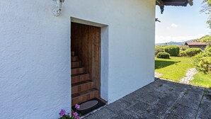 Ein weißes Gebäude mit einer Holztreppe, die zu einer Tür und einer Fußmatte führt, mit freiem Blick auf eine grüne Landschaft, Bäume und ein fernes Gebirge.