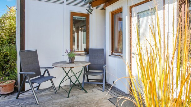 A wooden deck with two chairs and a table, a potted plant, and a window with blinds. - Holiday Apartment "Oestreich" close to Chiemsee Lake with Wi-Fi & Terrace (Chieming/ Egerer)