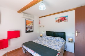A bedroom with a bed, a red chair, a wall-mounted air conditioner, a painting, a shelf with decorative items, and a door.