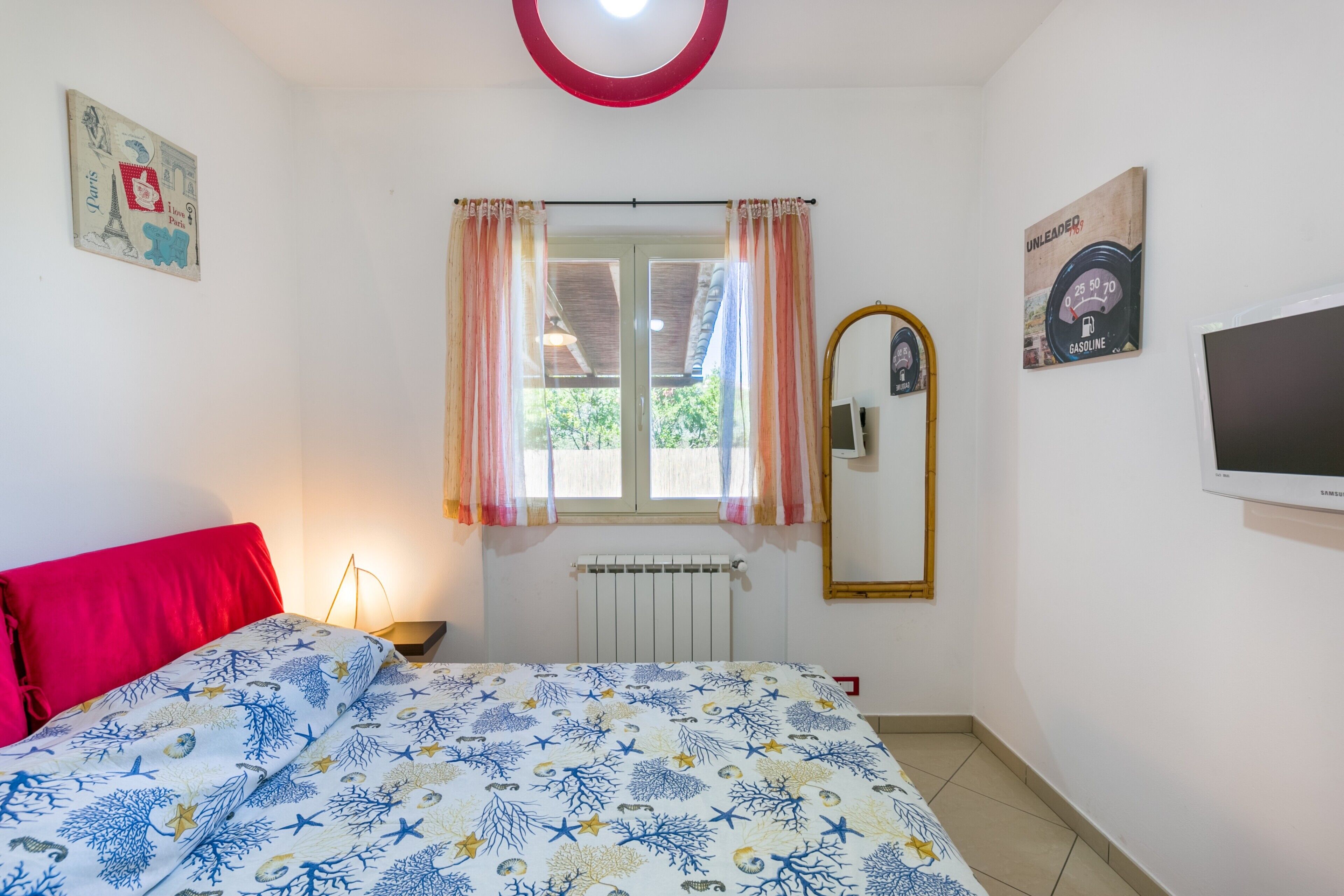 Un dormitorio con cama, cortinas en una ventana, un radiador, un espejo fijado a la pared y un televisor.