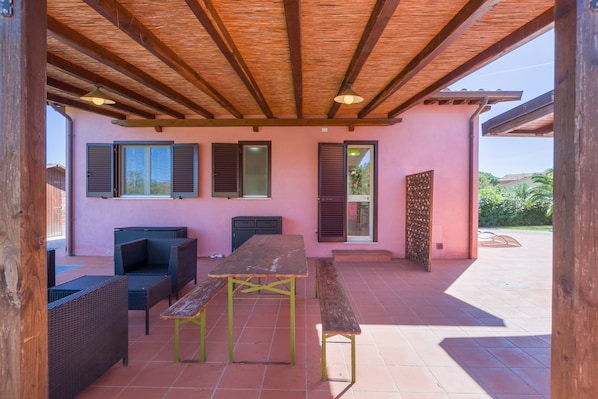 An outdoor patio with a wooden ceiling, a table, benches, wicker furniture, and a pink wall with windows and shutters.