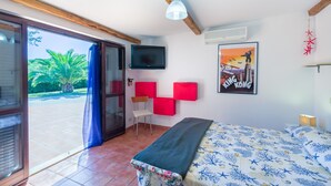 Un dormitorio con cama, televisor montado en la pared, una silla y puerta que da a un espacio exterior con vistas a zonas verdes.