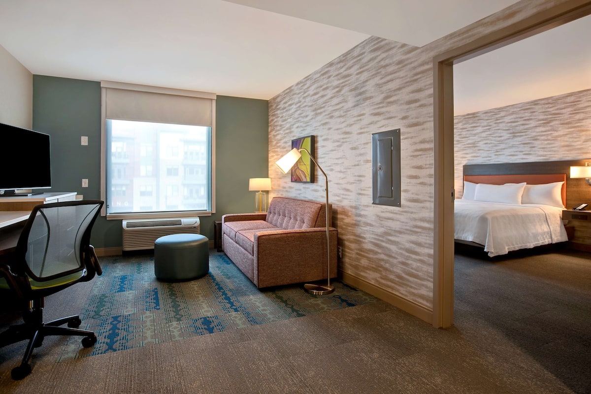 Une chambre d’hôtel avec un lit, un bureau équipé d’un ordinateur, une chaise, une lampe et une vue sur les bâtiments par la fenêtre.