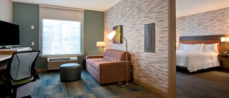 A hotel room with a bed, a desk with a computer, a chair, a lamp, and a view of buildings through the window.