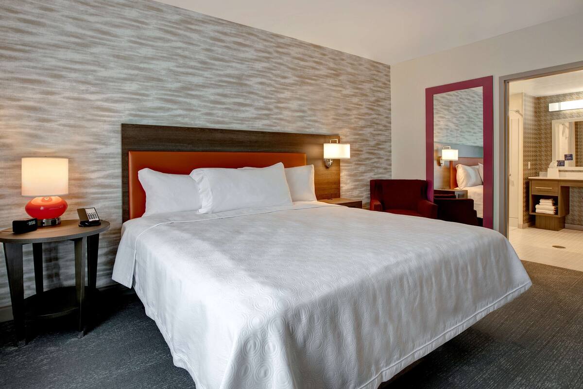 Une chambre d’hôtel avec un grand lit, une table de chevet avec une lampe, un fauteuil rouge et une salle de bain visible à travers une porte ouverte.