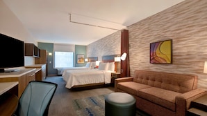 A hotel room with a bed, a sofa, a desk, a television, and a window. - Home2 Suites by Hilton Carmel Indianapolis (Carmel)