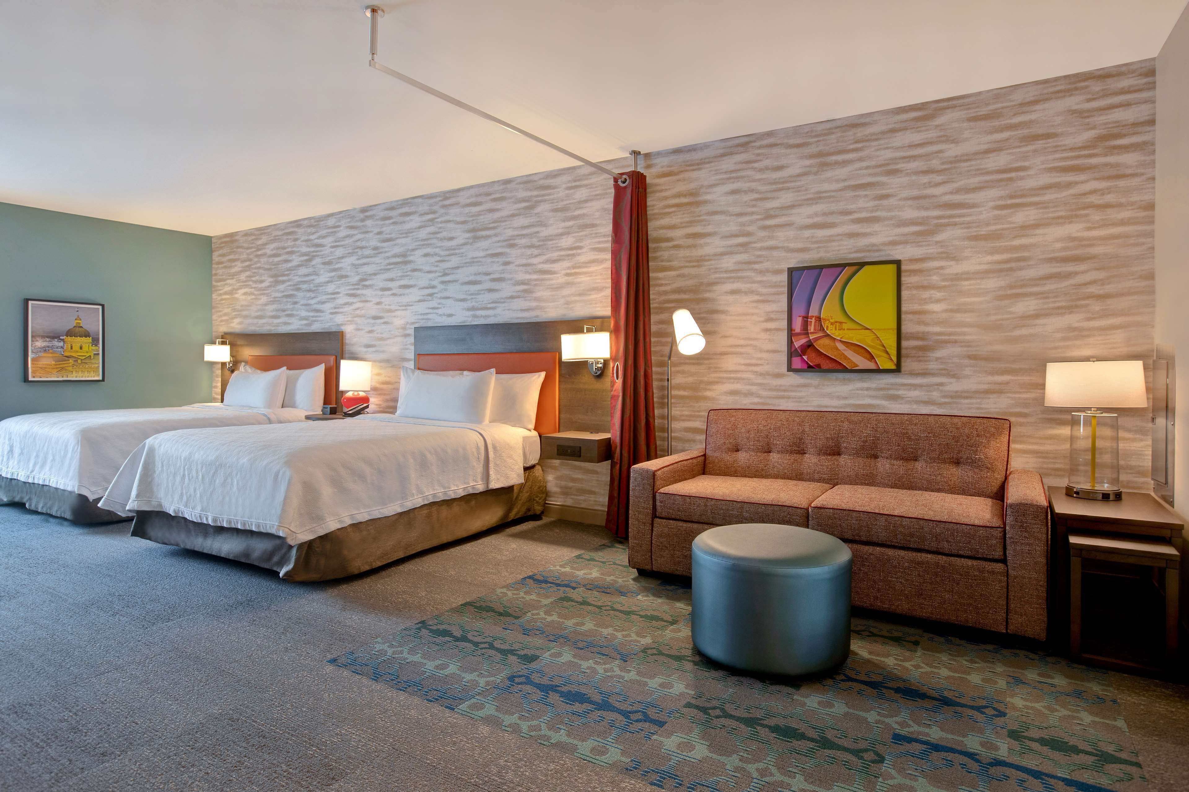 A hotel room with two beds, a sofa, a round ottoman, and a patterned rug.