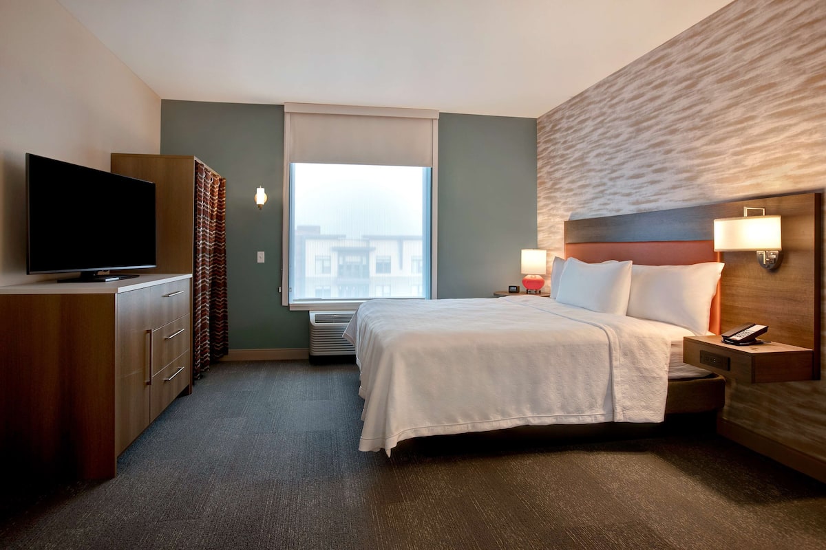Une chambre d’hôtel avec un grand lit, un téléviseur posé sur une commode en bois, une fenêtre donnant sur des immeubles et une lampe fixée au mur.