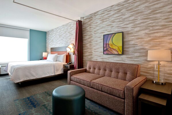 Quarto de hotel com cama, sofá, mesinha, luminária e um quadro na parede.