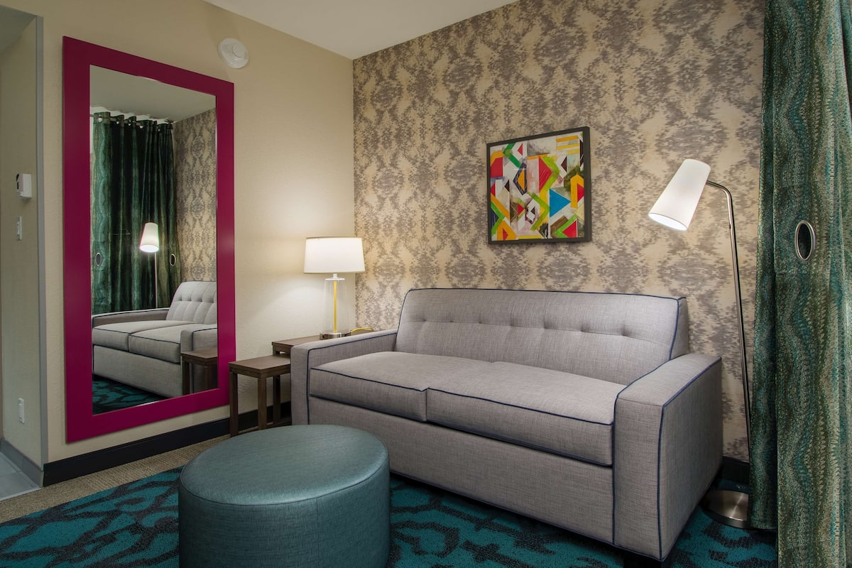 Een hotelkamer met een grijze bank, een rond poef, een spiegel met een roze lijst, een lamp en een kleurrijk abstract schilderij aan de muur.