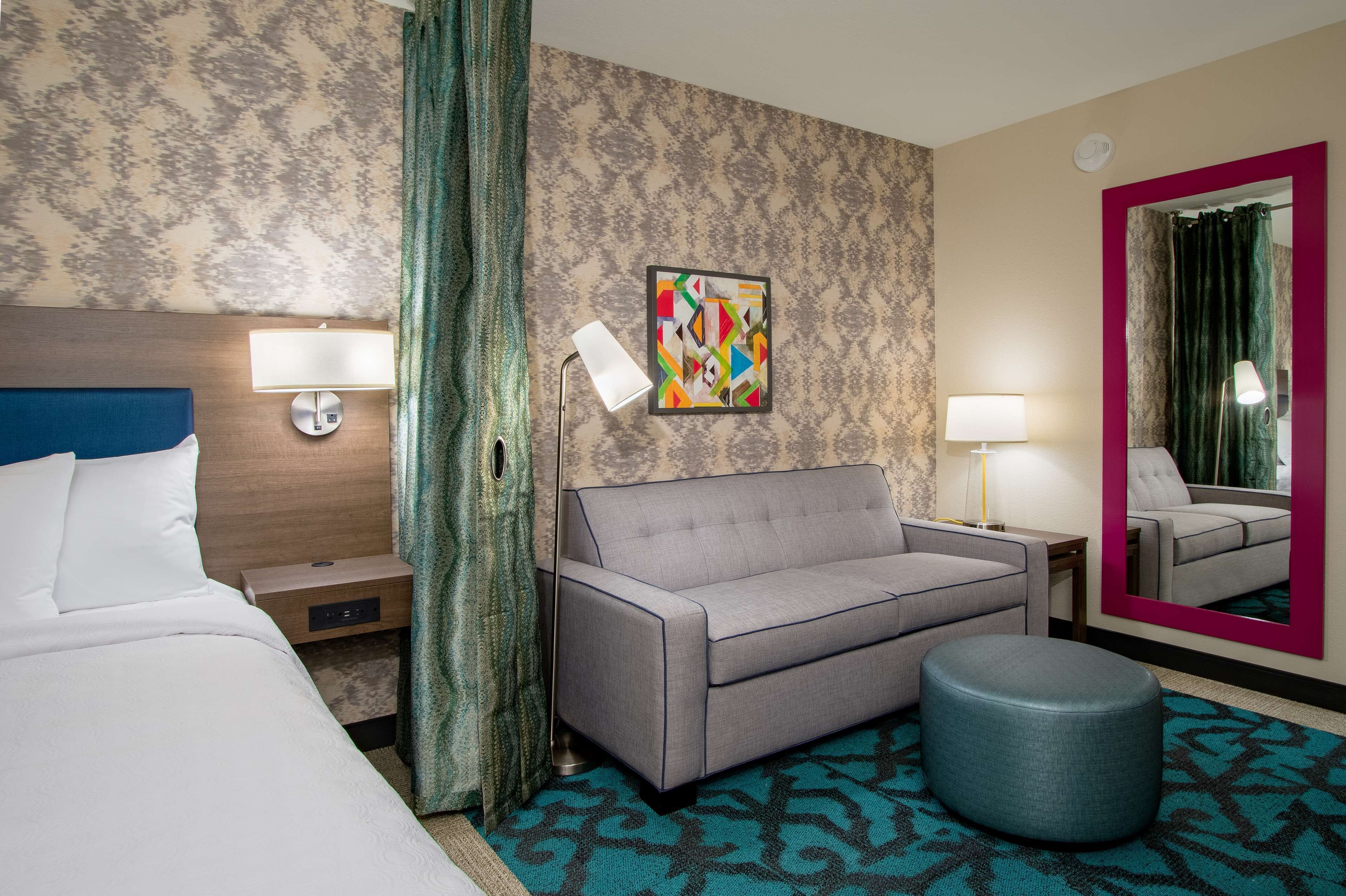 A hotel room with a bed, a sofa, a small ottoman, a wall-mounted lamp, a mirror, and a colorful artwork on the wall.