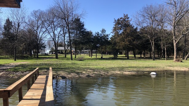 A wooden deck extending over a calm pond, surrounded by trees and grassy areas.