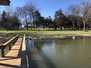 A wooden deck extending over a calm pond, surrounded by trees and grassy areas.