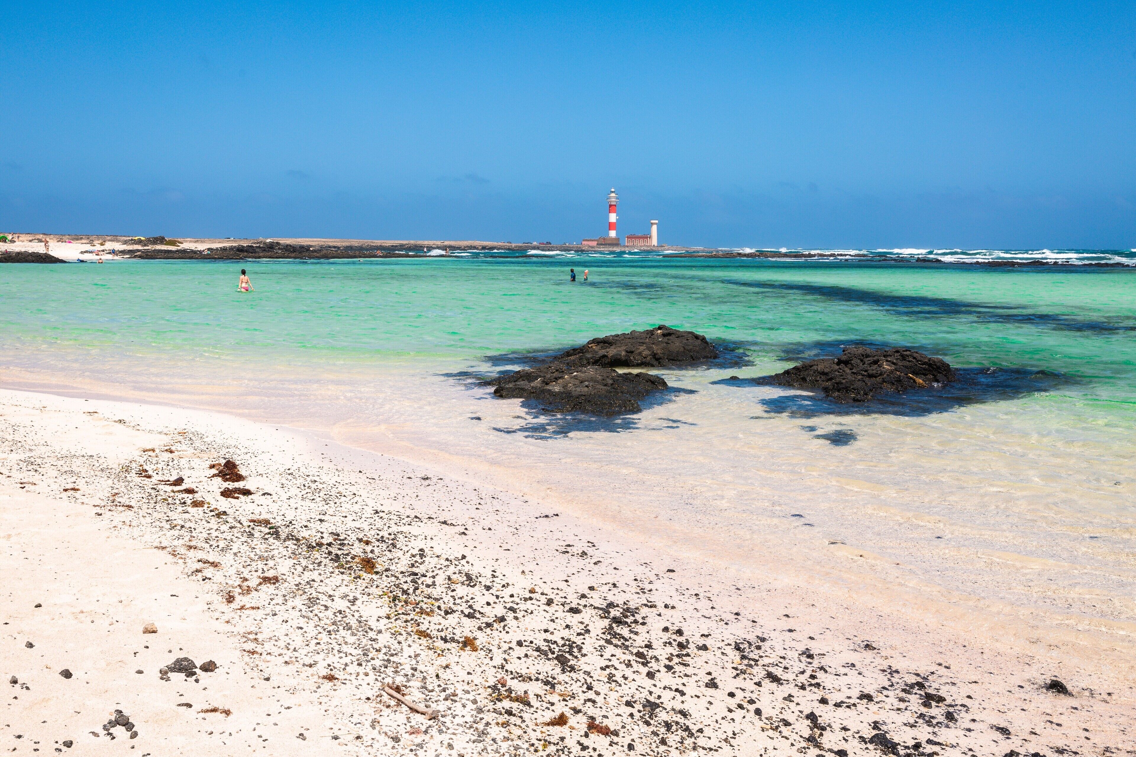 Ein Strand mit klarem türkisblauem Wasser, ein Leuchtturm in der Ferne und Felsformationen.