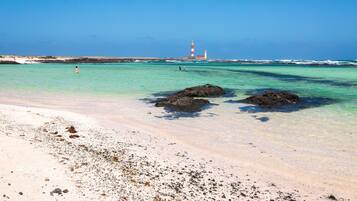 Ein Strand mit klarem türkisblauem Wasser, ein Leuchtturm in der Ferne und Felsformationen.
