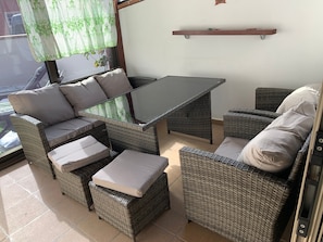 Un patio ensoleillé, aménagé avec des meubles en osier, notamment une table et des chaises, et une petite étagère fixée au mur.