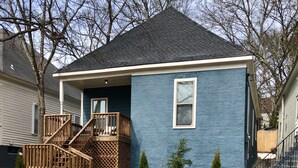 Una casa de dos pisos con fachada azul, un porche delantero de madera con escalera y un muro de piedra al frente.