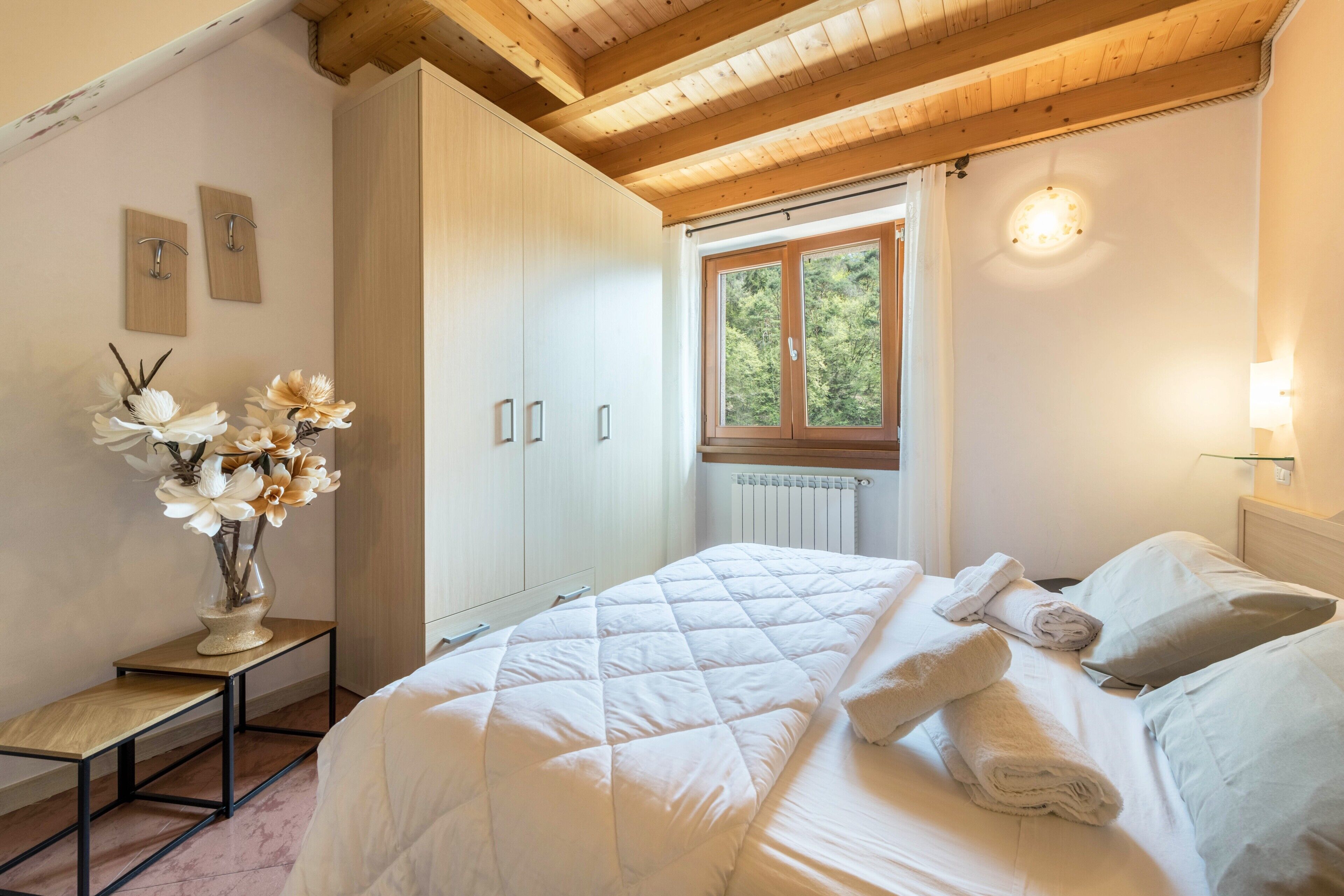 A bedroom with a bed, a nightstand with a vase of flowers, a window with a view of trees, and a wooden ceiling.
