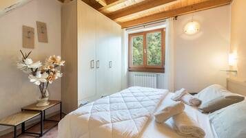 A bedroom with a bed, a nightstand with a vase of flowers, a window with a view of trees, and a wooden ceiling.