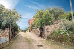 Un sentier rural mène à un bâtiment aux murs de pierre, doté d’un portail en métal, et entouré de verdure sous un ciel dégagé.