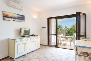 A room with a TV, a cabinet, a balcony with a table and chairs, and a painting on the wall. - Apartment 'Giardini Di Bados B3' with Shared Terrace, Shared Garden and Air Conditioning (pittulongu)