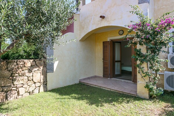 A building with a stone wall, a small grassy area, and a doorway with wooden shutters.