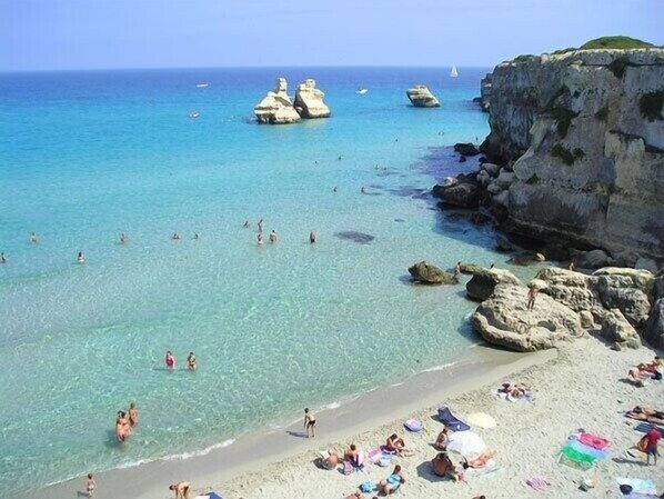 Une plage avec une eau cristalline, des formations rocheuses, et des gens qui se baignent et prennent des bains de soleil.
