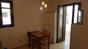 Une pièce avec une table et des chaises en bois, un téléviseur posé sur un meuble, et une porte-fenêtre coulissante donnant sur un espace extérieur.