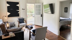A cozy living room with a sofa, armchair, and coffee table. A large abstract painting adorns the wall, and a television is mounted above a door. - NEW! Hip Downtown Pad! Walk Everywhere! (Hot Springs)