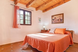 A bedroom with a wooden ceiling, a bed with an orange bedspread, a nightstand with a lamp, and a window with a curtain.