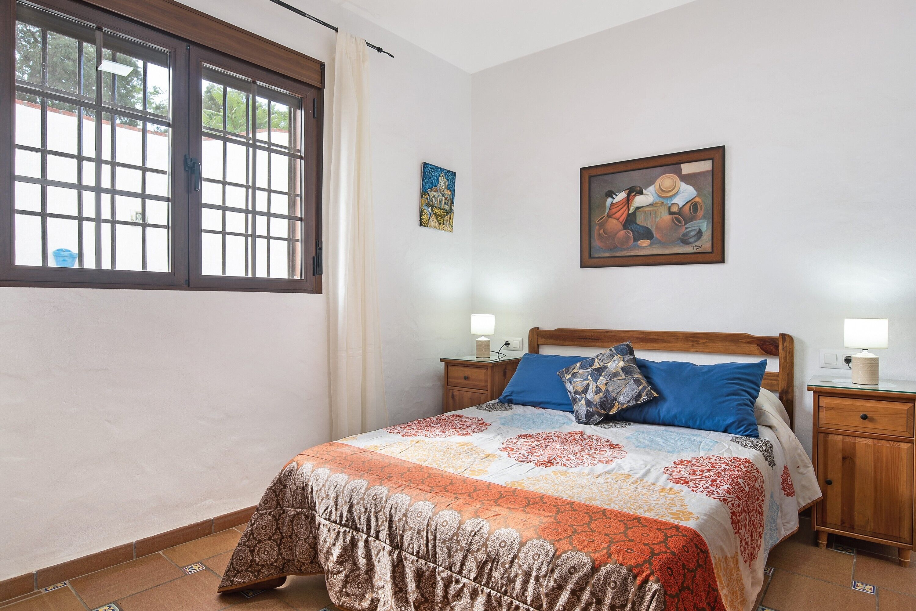 A bedroom with a large window, a bed with patterned bedding, two bedside tables with lamps, and a painting on the wall.