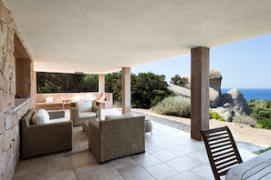 A covered outdoor seating area with wicker furniture, a stone wall, and a view of the sea and rocky landscape.