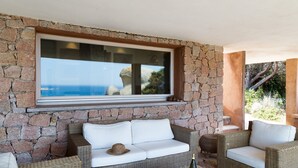 Un muro de piedra con un amplio ventanal que da a un paisaje, muebles de mimbre y una botella sobre una mesa.