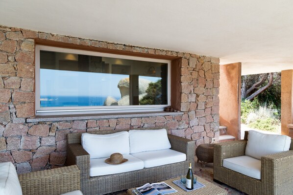 A stone wall with a large window overlooking a landscape, wicker furniture, and a bottle on a table.