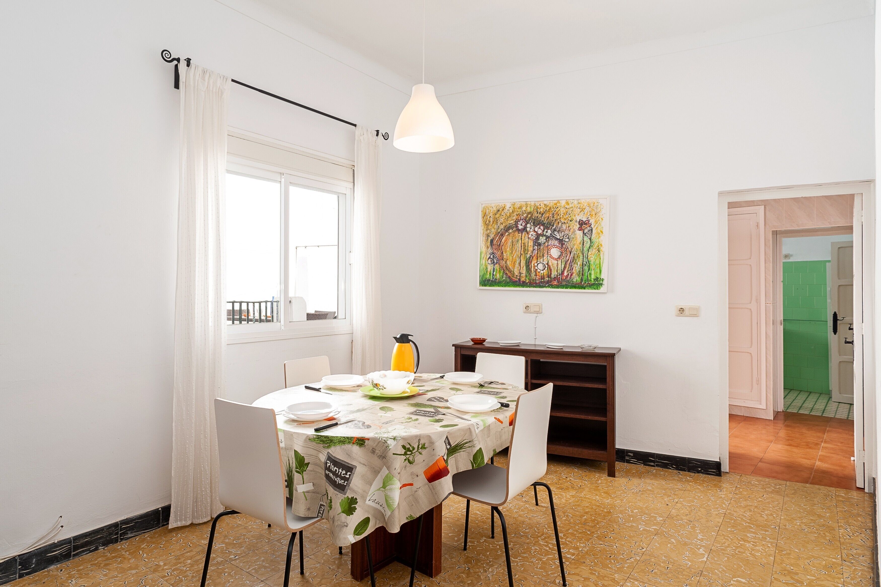 A dining area with a table set for four, a painting on the wall, and a view of the outside through a window.