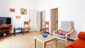 Un salon comprenant un téléviseur, deux fauteuils, une table avec un saladier, et une porte donnant sur une autre pièce.