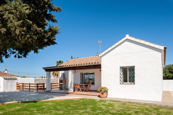 A white building with a tiled roof, a patio with a table and chairs, and a potted plant.
