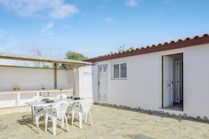 Une terrasse avec une table et des chaises, un bâtiment blanc avec un toit en tuiles et une porte donnant sur un espace intérieur.