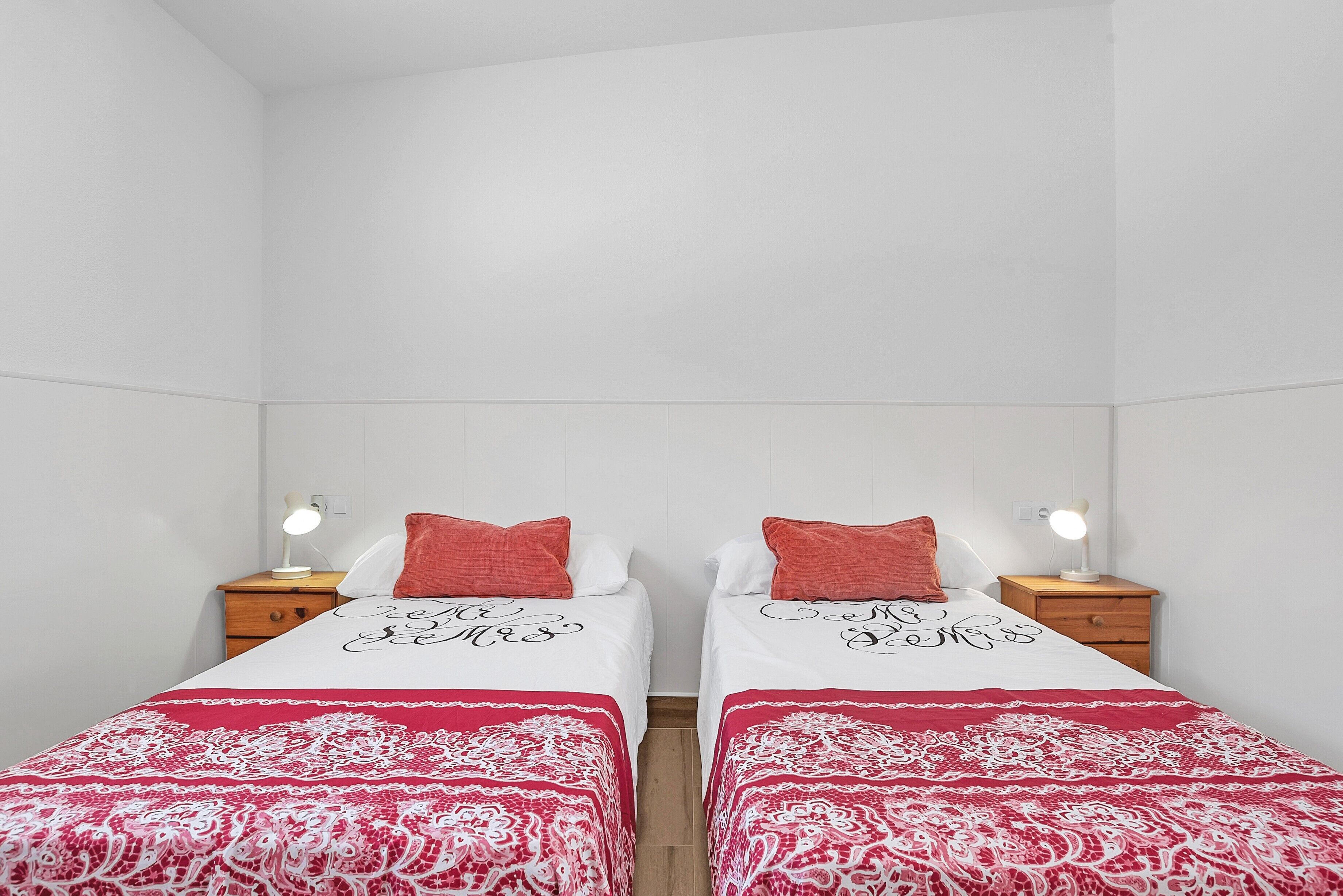 A twin room with two beds, each with a red and white patterned bedspread, two bedside tables with lamps, and a white tiled wall.