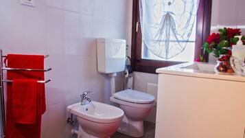 Ein Badezimmer mit zwei Toiletten, einem Bidet, einem Waschbecken und einem Fenster mit einem Zierrollo.