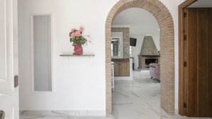 Un couloir avec une porte blanche, un vase rose orné de fleurs, une arche en briques, un coin cuisine avec une cheminée en pierre et un canapé rose.