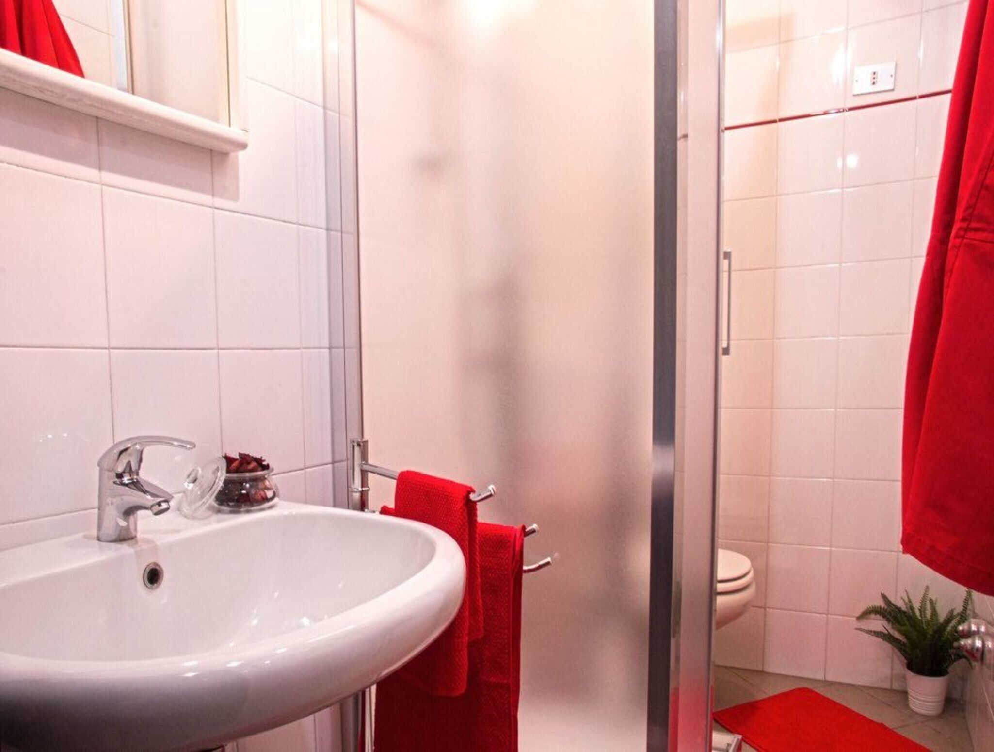 Un bagno con un lavandino bianco, asciugamani rossi, box doccia in vetro e un water.