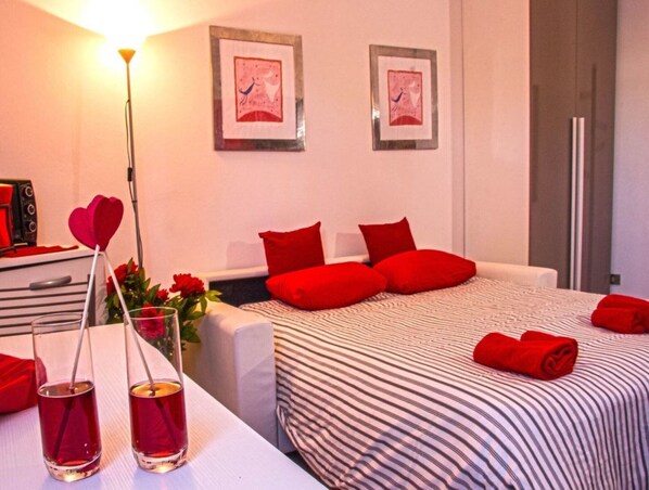 Une chambre d’hôtel avec un lit, deux oreillers rouges, une table de chevet avec une lampe, et deux tableaux encadrés accrochés au mur.