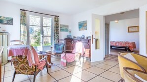 A spacious living room with a large window, a dining area with a table set for four, and a cozy seating area with chairs and a sofa. - Sunny Lodge by Interhome (Cabourg)