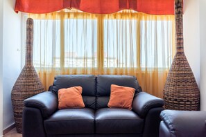 A black leather sofa with two orange cushions, a large window with sheer curtains, and two tall, woven vases on either side.