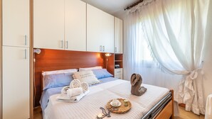 Ein ordentlich bezogenes Bett mit Kissen, ein Nachttisch mit Lampe und ein Fenster mit durchscheinenden Vorhängen.