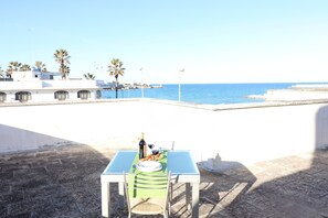 A table with a bottle, glasses, and food on a terrace overlooking the sea.