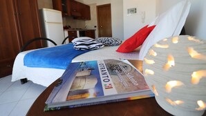Un dormitorio con cama, almohadas, una revista, una lámpara y una cocina al fondo.