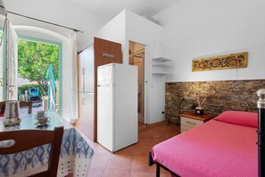 Ein kleines Zimmer mit einem Bett, einem Tisch mit Stuhl, einem Kühlschrank und einer Steinwand.